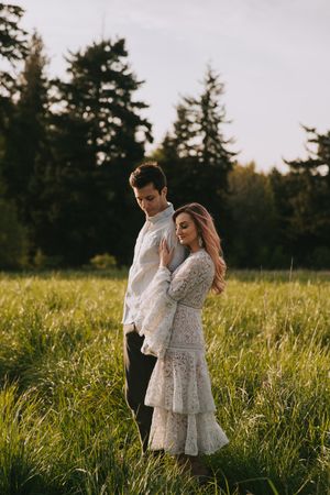 A couple stands back-to-back in a grassy field at golden hour with evergreen trees in the background.