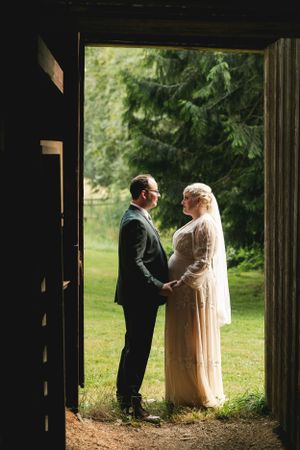 A romantic wedding couple standing in a doorway framed by evergreen trees and natural light.