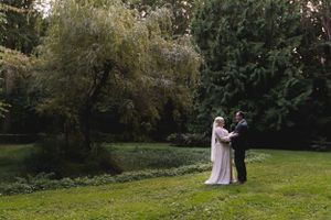 A wedding couple embracing on a lush green lawn surrounded by willow trees.