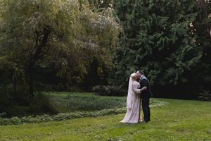 A tender moment between newlyweds dancing on a serene garden lawn beneath willow trees.