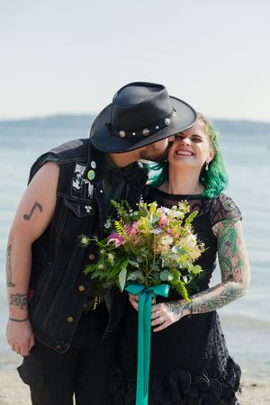 A couple in alternative wedding attire poses with a colorful bouquet against an oceanfront backdrop.