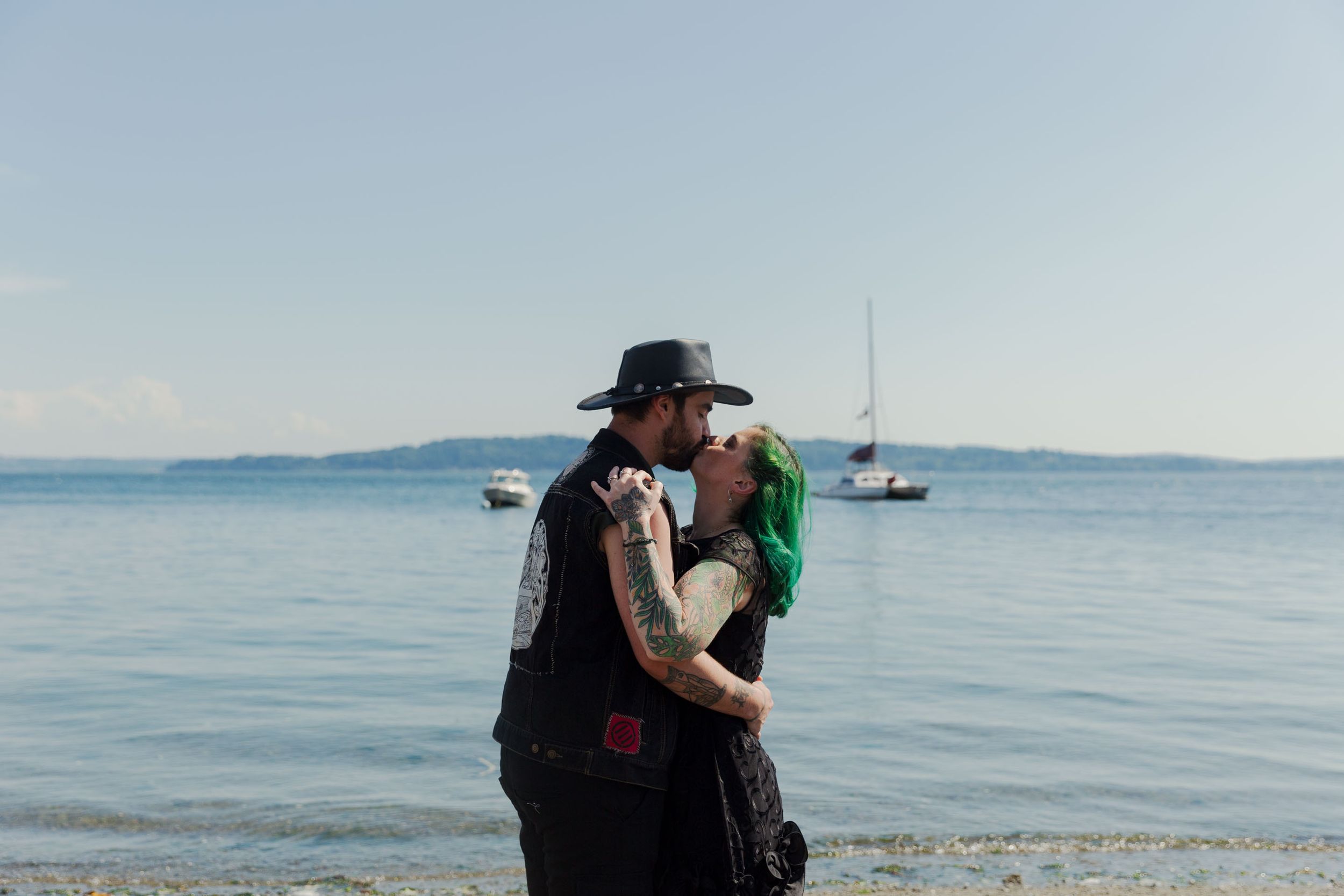 A couple embraces tenderly by the water with boats visible in the distance.