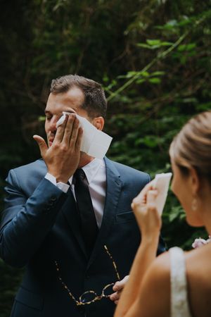 Couple sharing an emotional first look moment at wedding ceremony while wiping tears with tissues.
