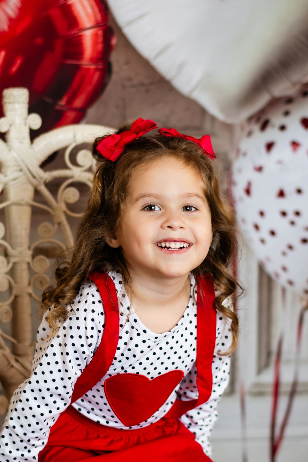 A young child in a polka dot shirt and red overalls with bows smiles brightly against a decorated holiday backdrop.