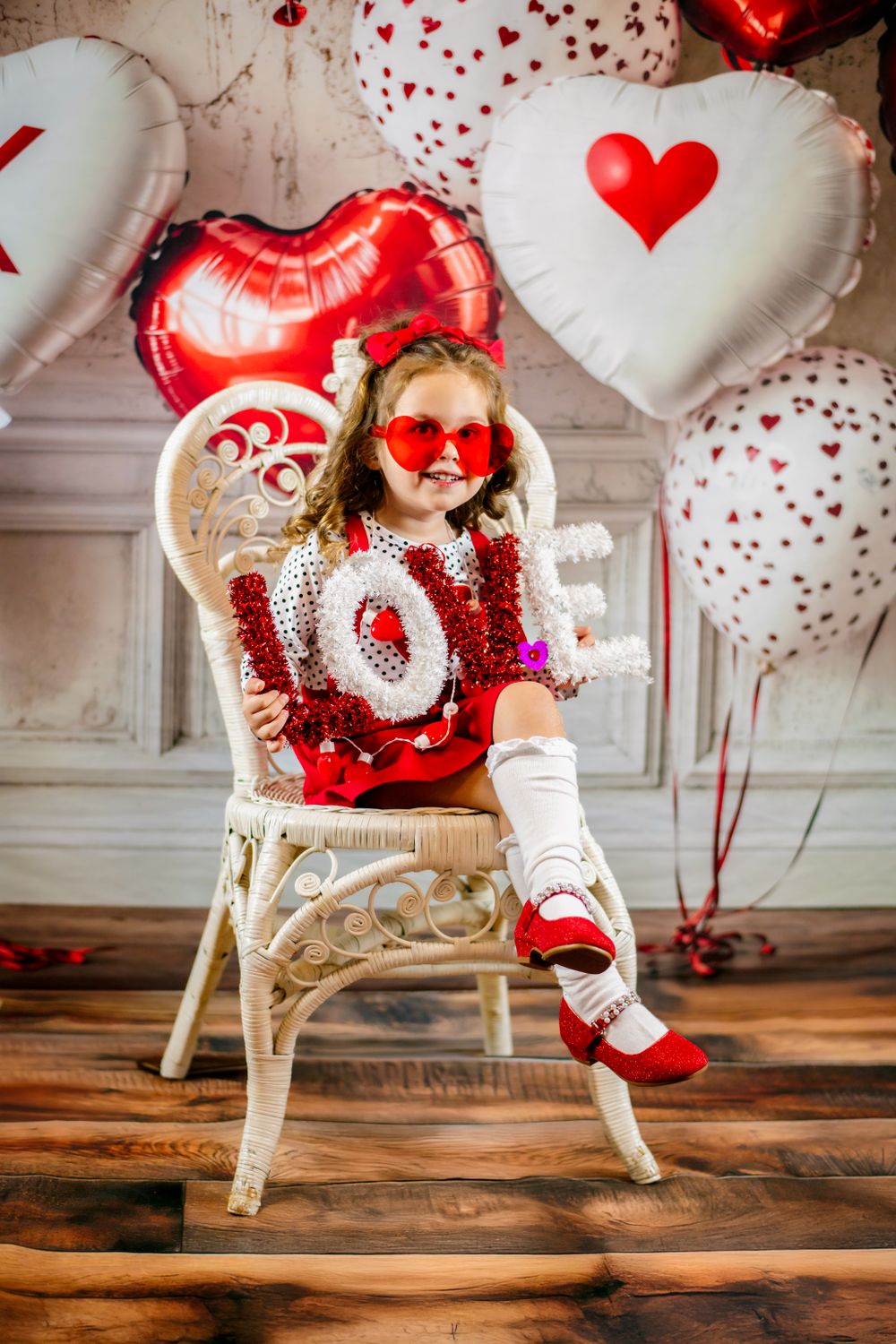 Valentine's Day decorative scene with heart-shaped balloons, wicker chair, and red and white festive decor on wooden floor.