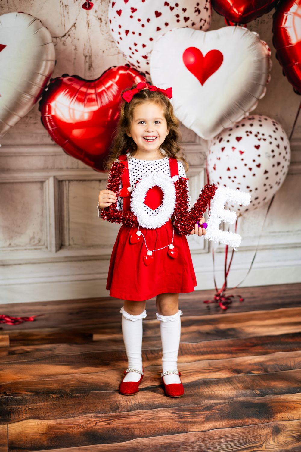 Valentine's Day photo setup with heart balloons, polka dot dress, knee socks and red shoes against grey backdrop.