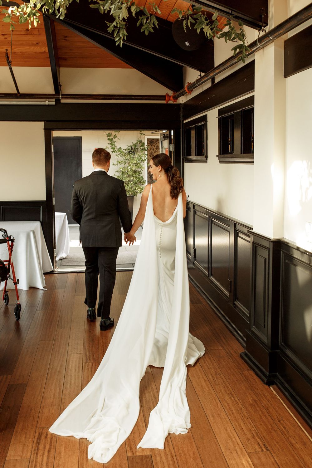 Elegant wedding couple walking down wooden hallway with flowing white dress train.