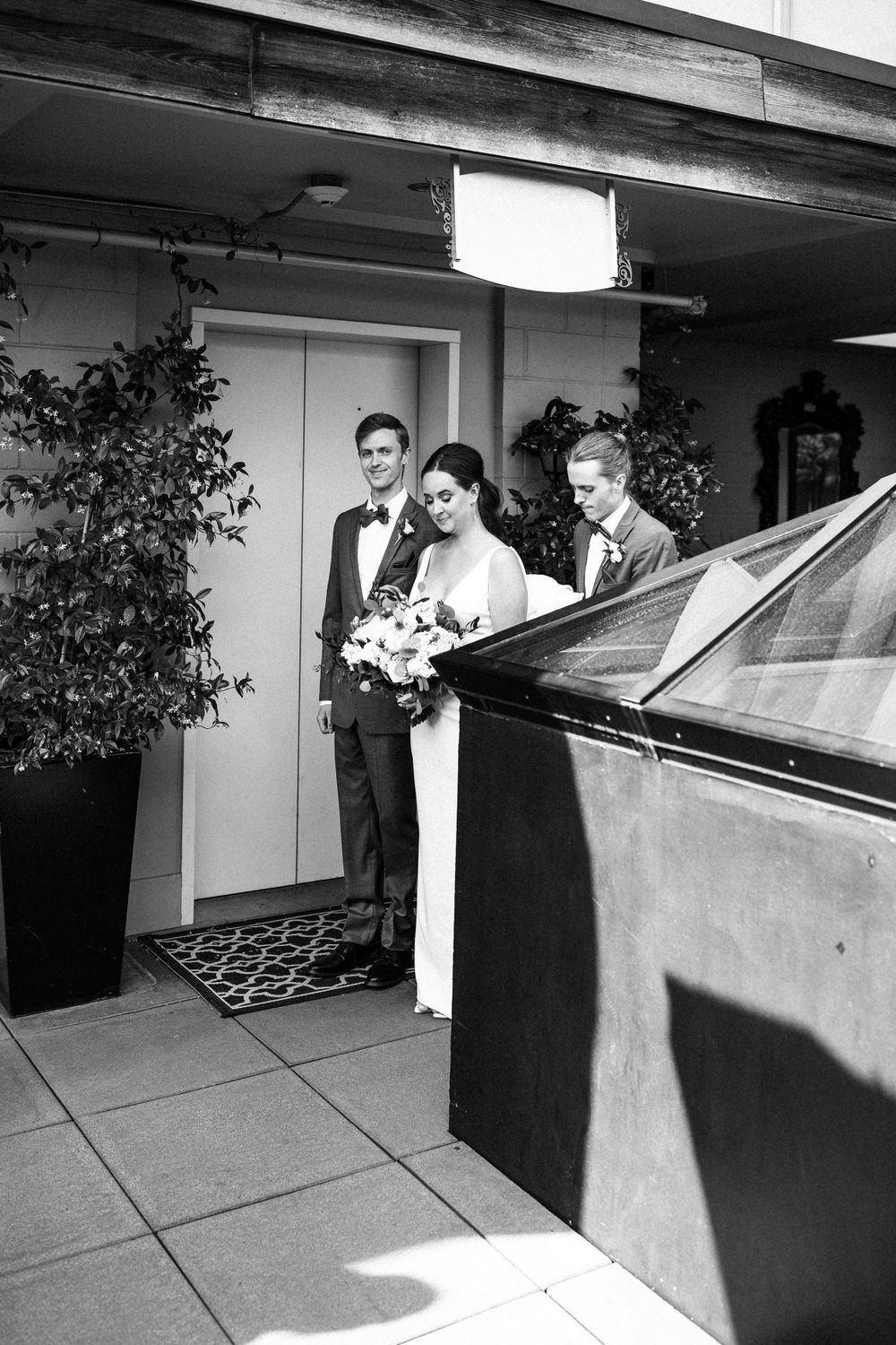 Wedding party members stand near modern building entrance in formal attire.
