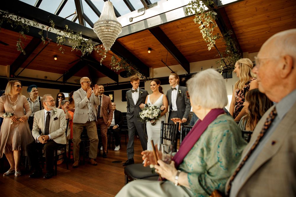 Wedding ceremony guests watch attentively from chairs in a rustic venue with wooden beams and floral decor.