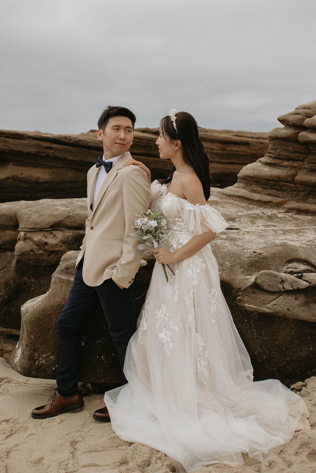 A couple in wedding attire sits together on coastal rocks during a moody overcast photoshoot at Windansea Beach in La Jolla.