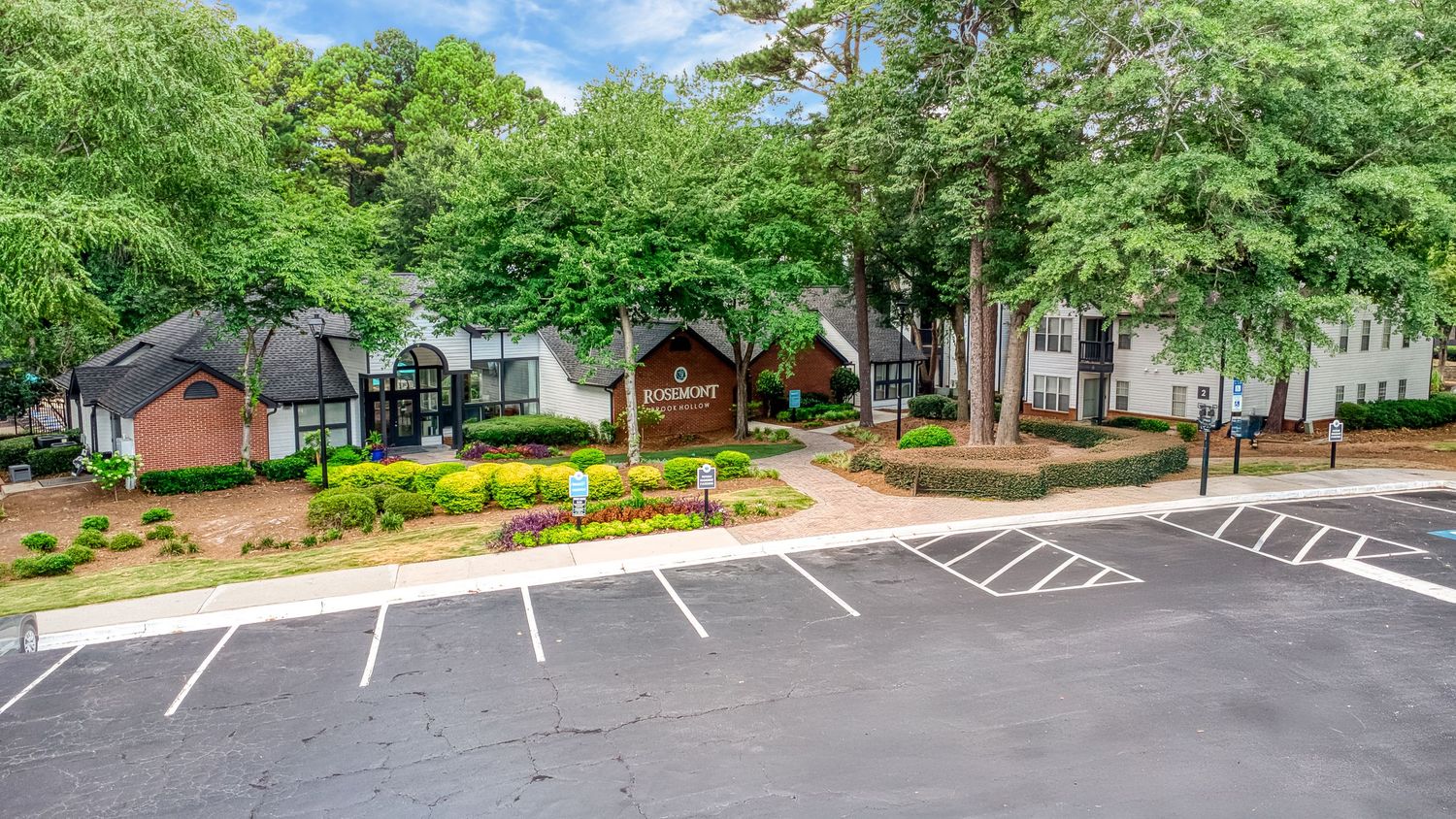 Panoramic view of a tree-lined parking area with empty spaces and brick buildings visible in the background.