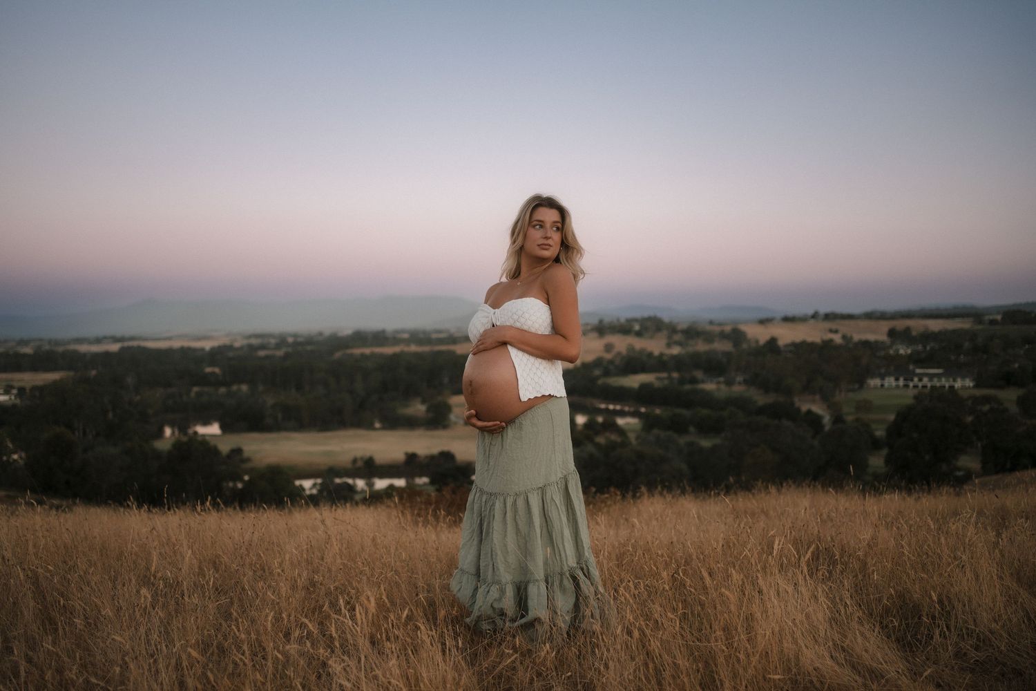 Pregnant woman in white top and sage skirt stands in golden field at sunset overlooking rural valley landscape.
