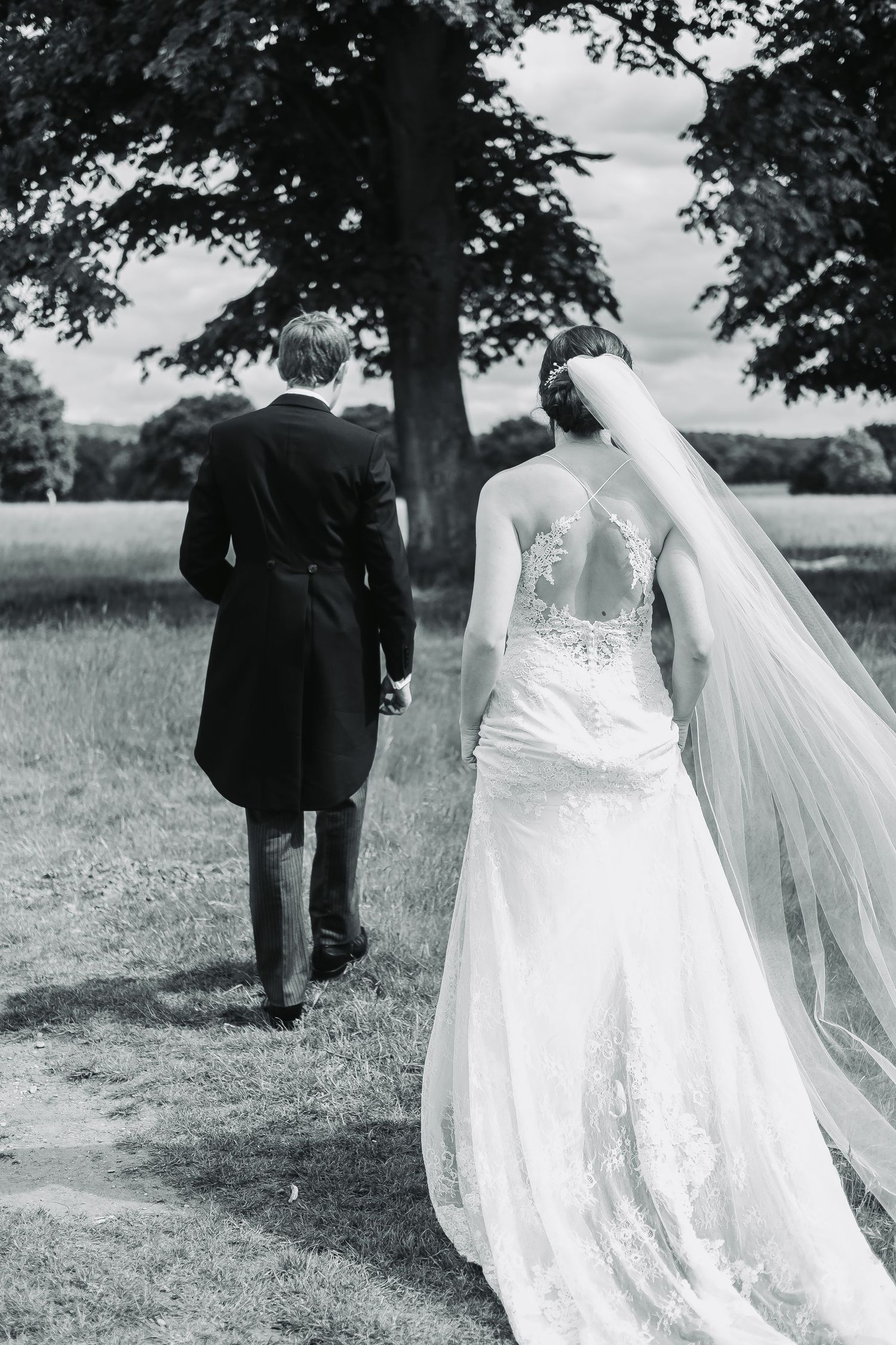 A black and white wedding photo showing a bride in a lace gown with flowing veil walking behind groom in formal attire outdoors.