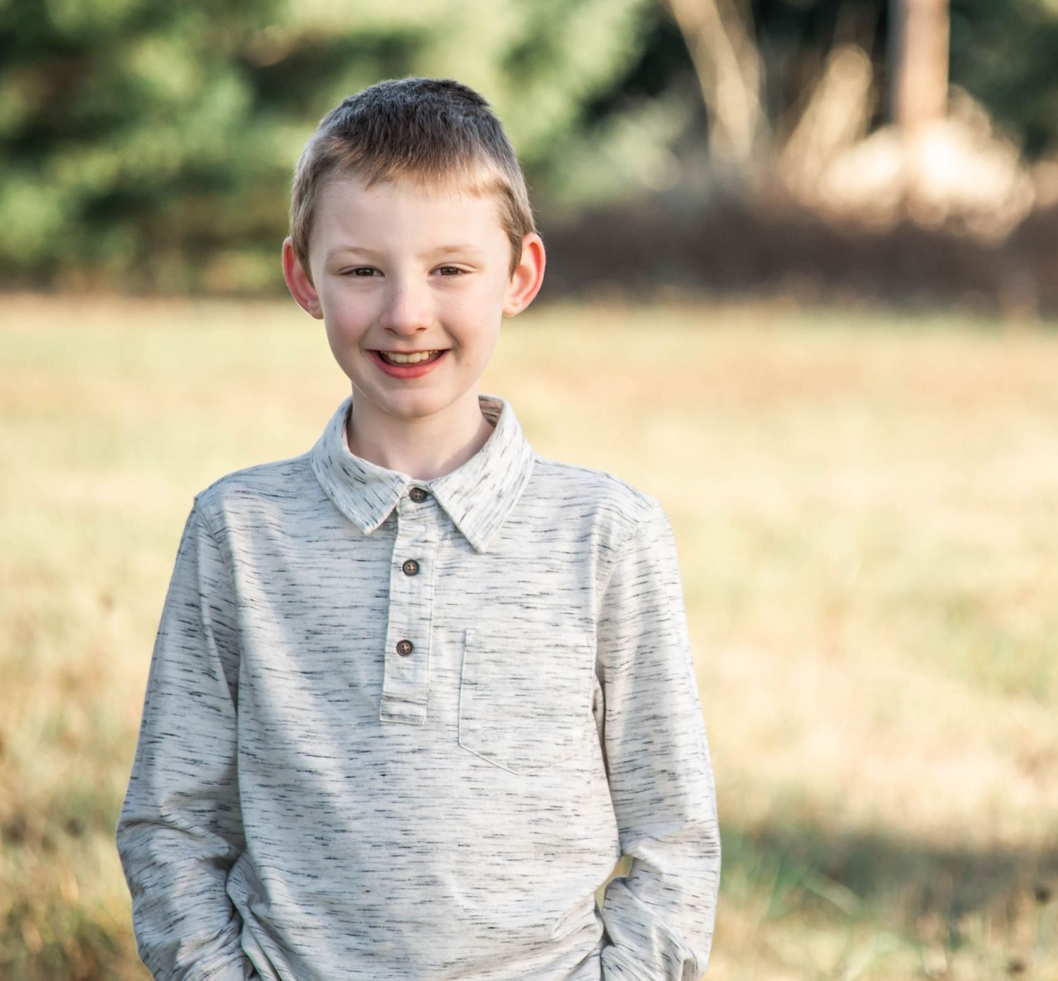 Young person wearing a gray polo shirt smiles warmly in an outdoor setting with natural lighting.