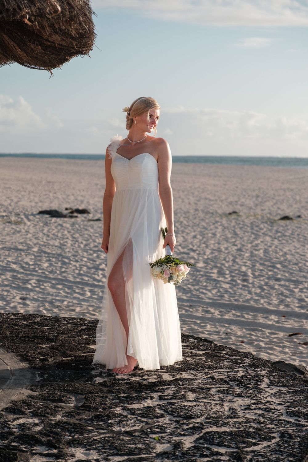 A bride in flowing white dress stands on a sandy beach with ocean views.