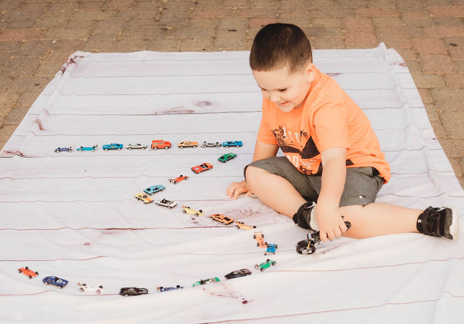 A child in an orange shirt plays with toy cars on a large white paper surface spread across the floor.