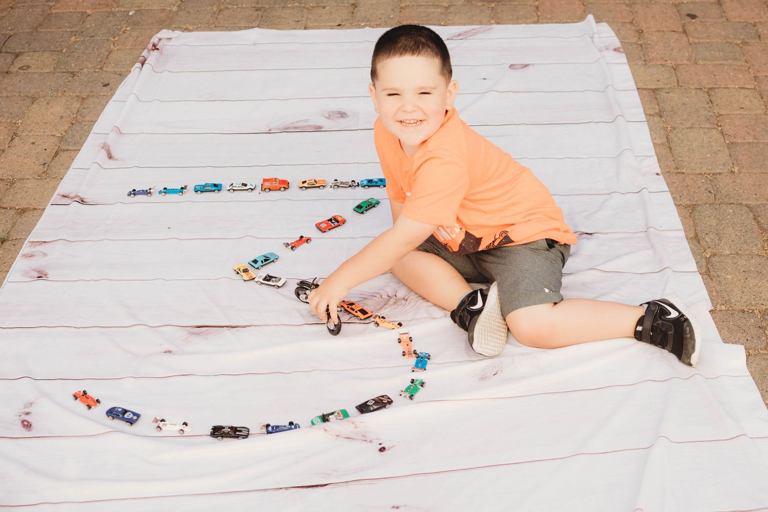 A person lays on a tiled floor playing with a long line of colorful toy cars and trucks.