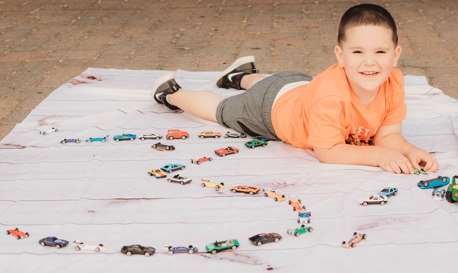 A child in an orange shirt lies on a blanket surrounded by toy cars arranged in a spiral pattern.