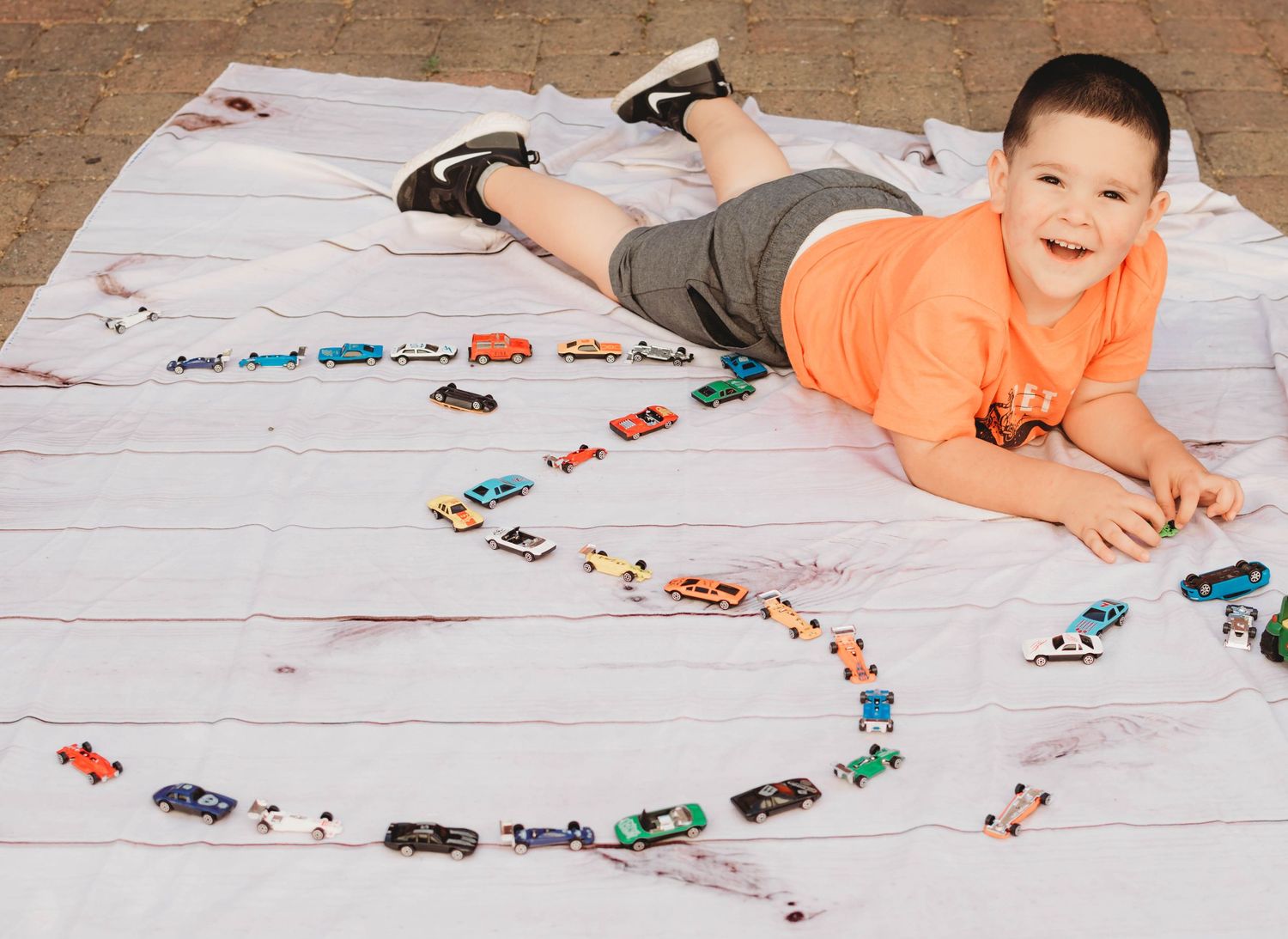 A long line of toy cars and vehicles arranged on a light colored floor with someone lying down beside them.