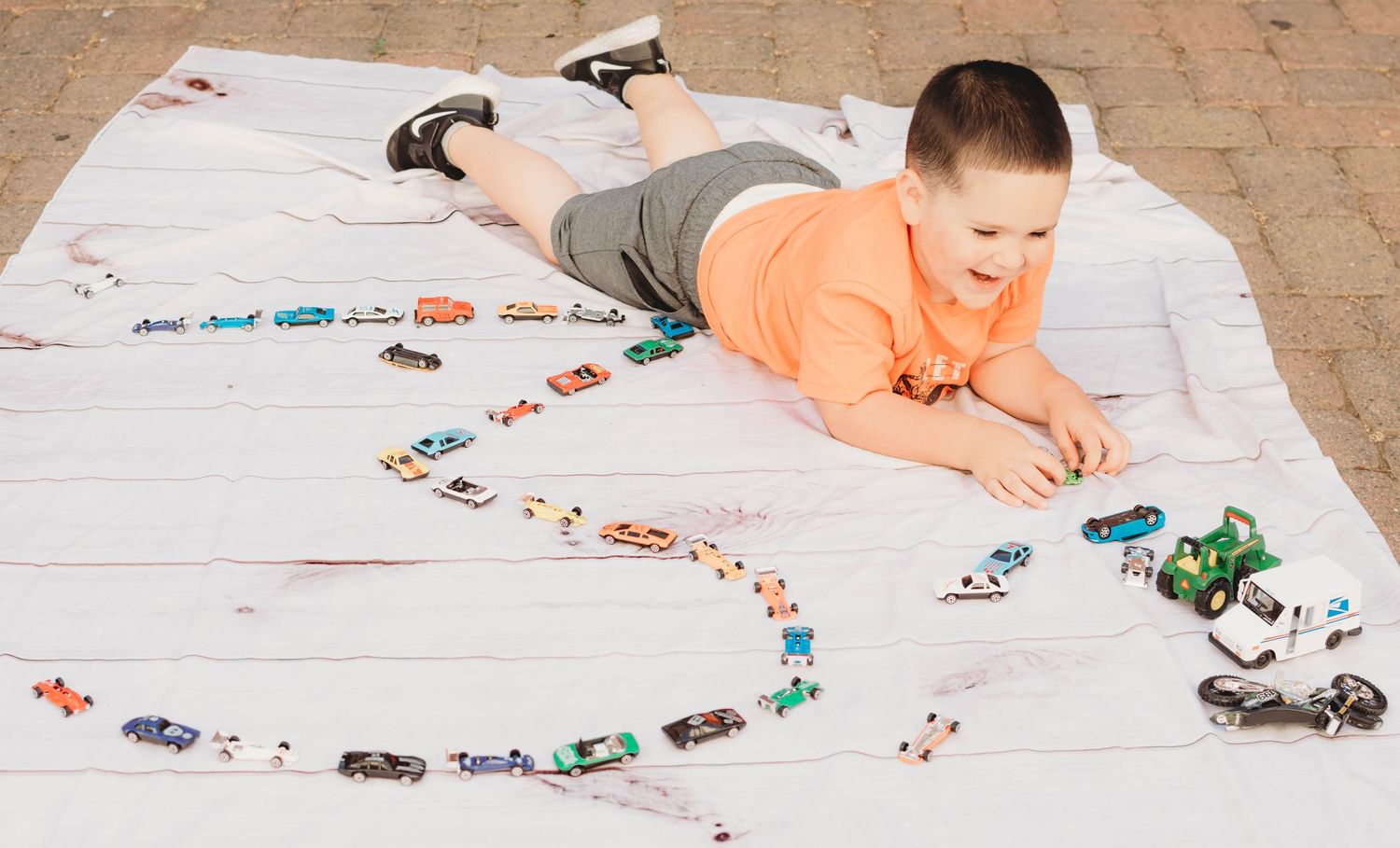 Child plays with toy cars and vehicles on a white blanket against a brick wall background.