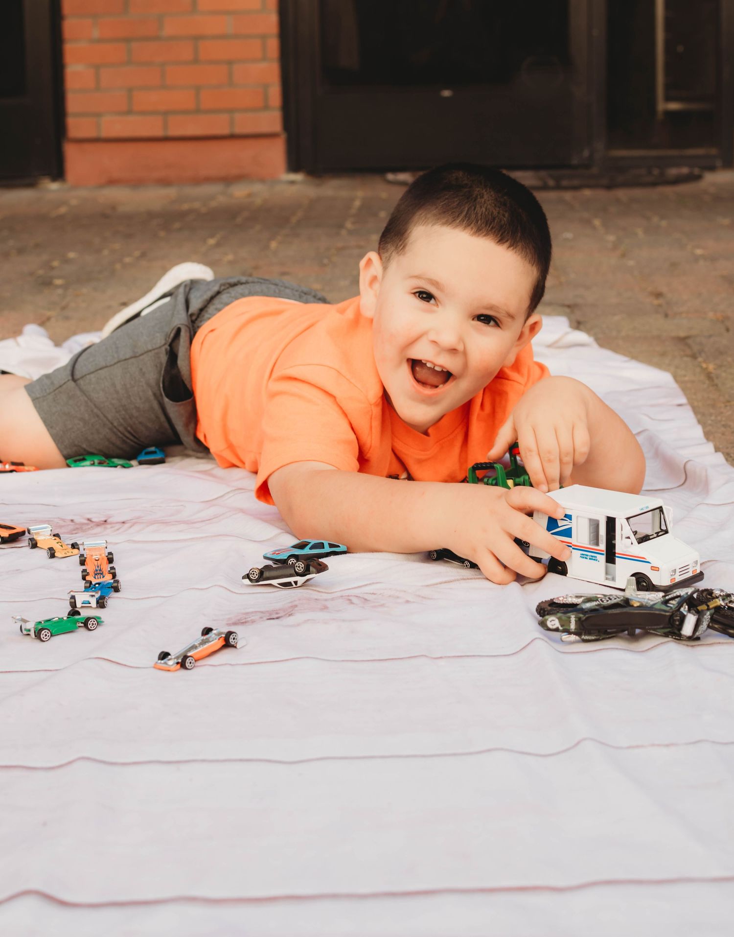 Child arranges colorful toy cars and trucks while lying on white blanket near brick building.
