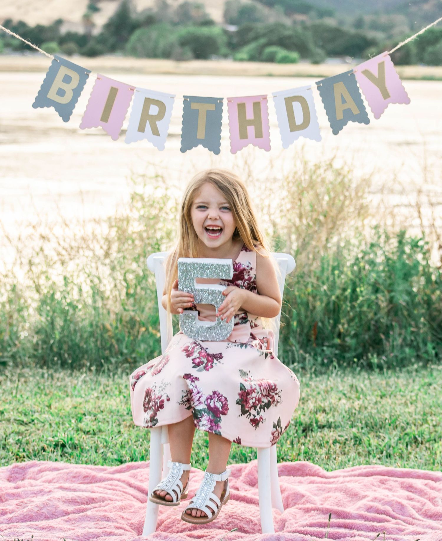 A birthday celebration scene with a pink blanket on grass and decorative bunting hanging overhead.