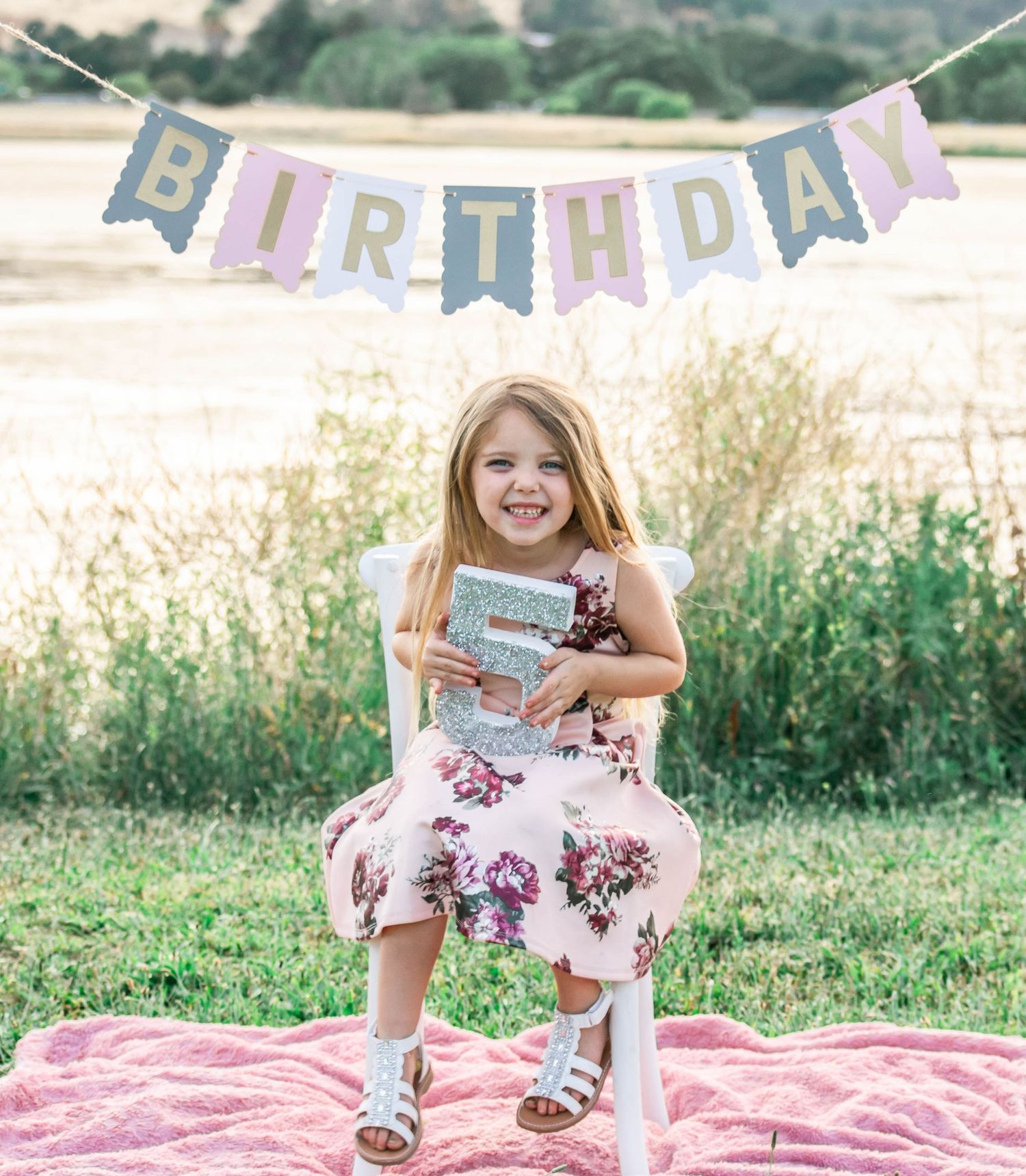 A birthday celebration setup outdoors with a decorative banner and pink blanket on grass near a lake.
