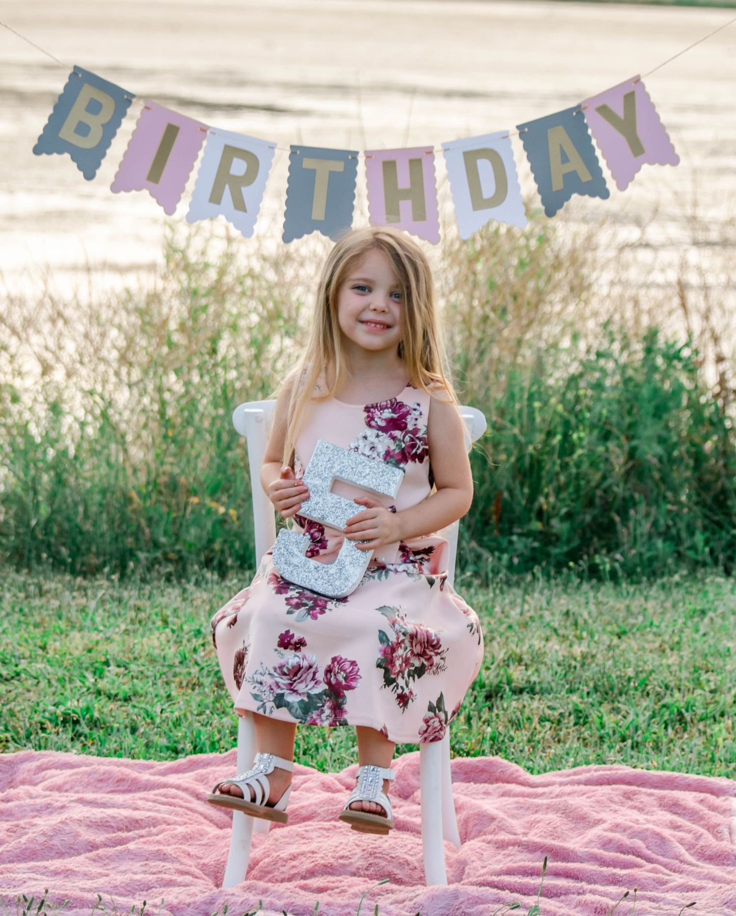 Birthday celebration setup on pink blanket outdoors with decorative bunting and party decor in natural setting.