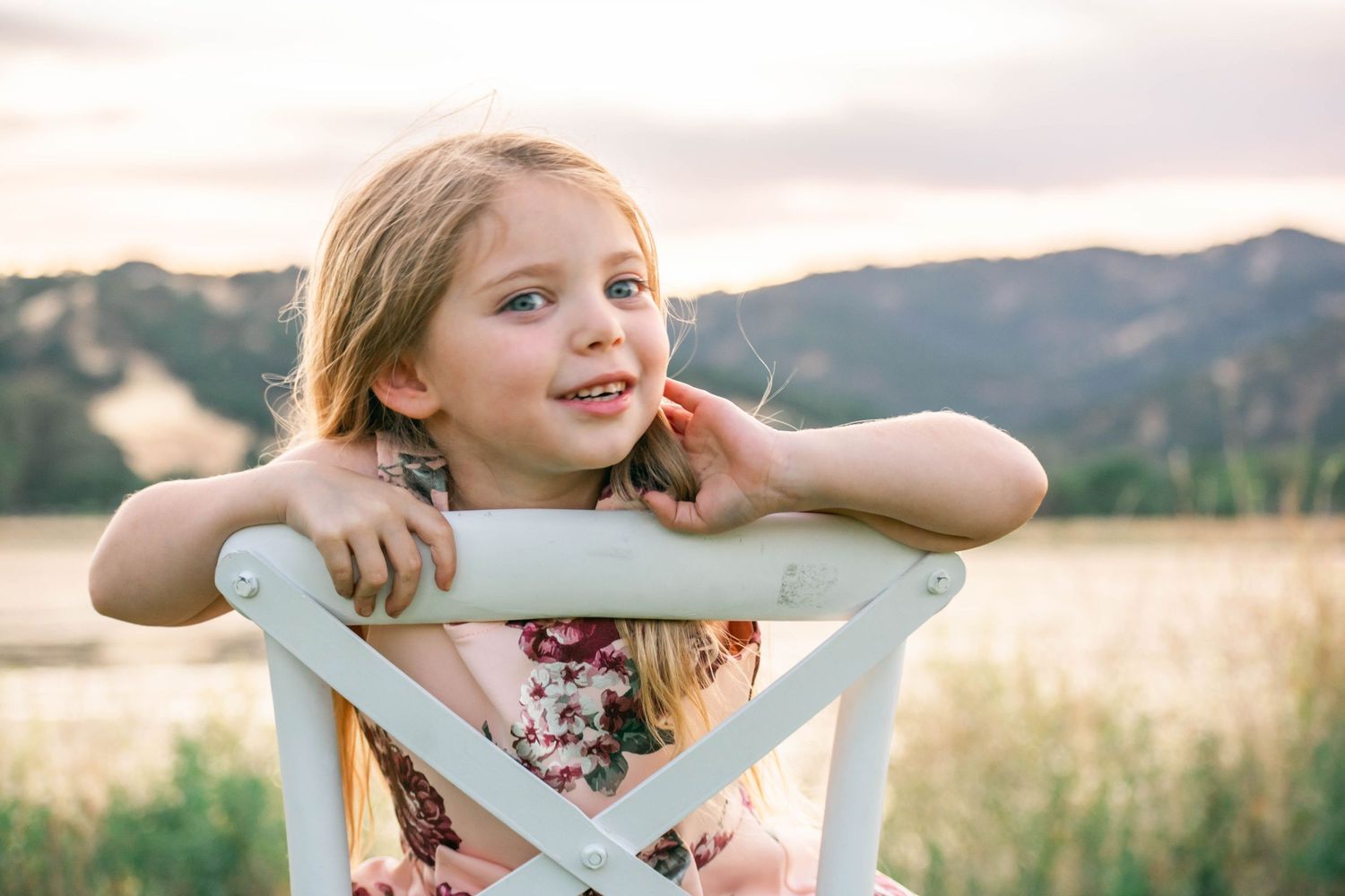 Child sitting in white wooden chair outdoors in field during golden hour sunset.