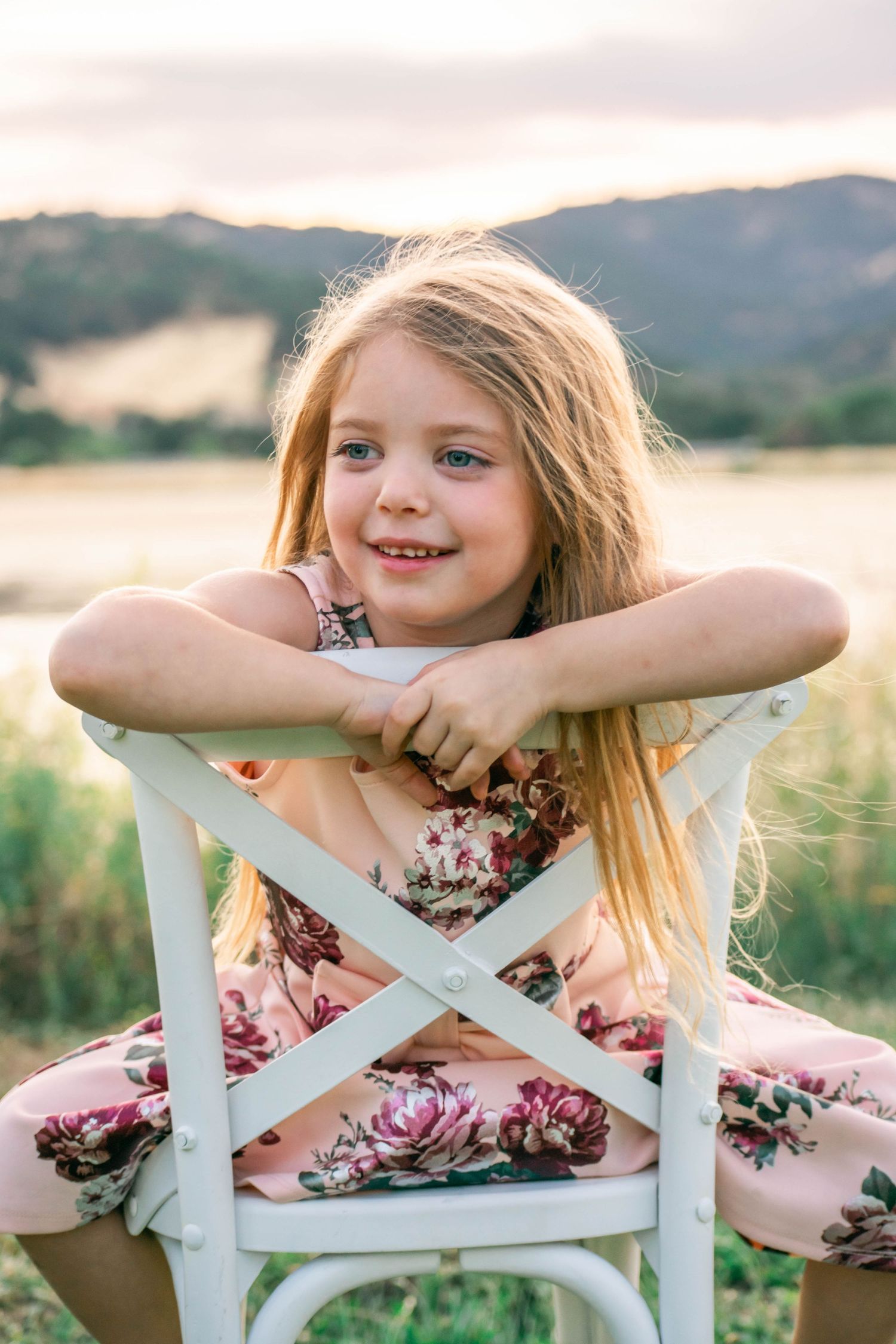 A child in a floral dress sits backwards on a white wooden chair in a sunlit meadow with mountains in the background.