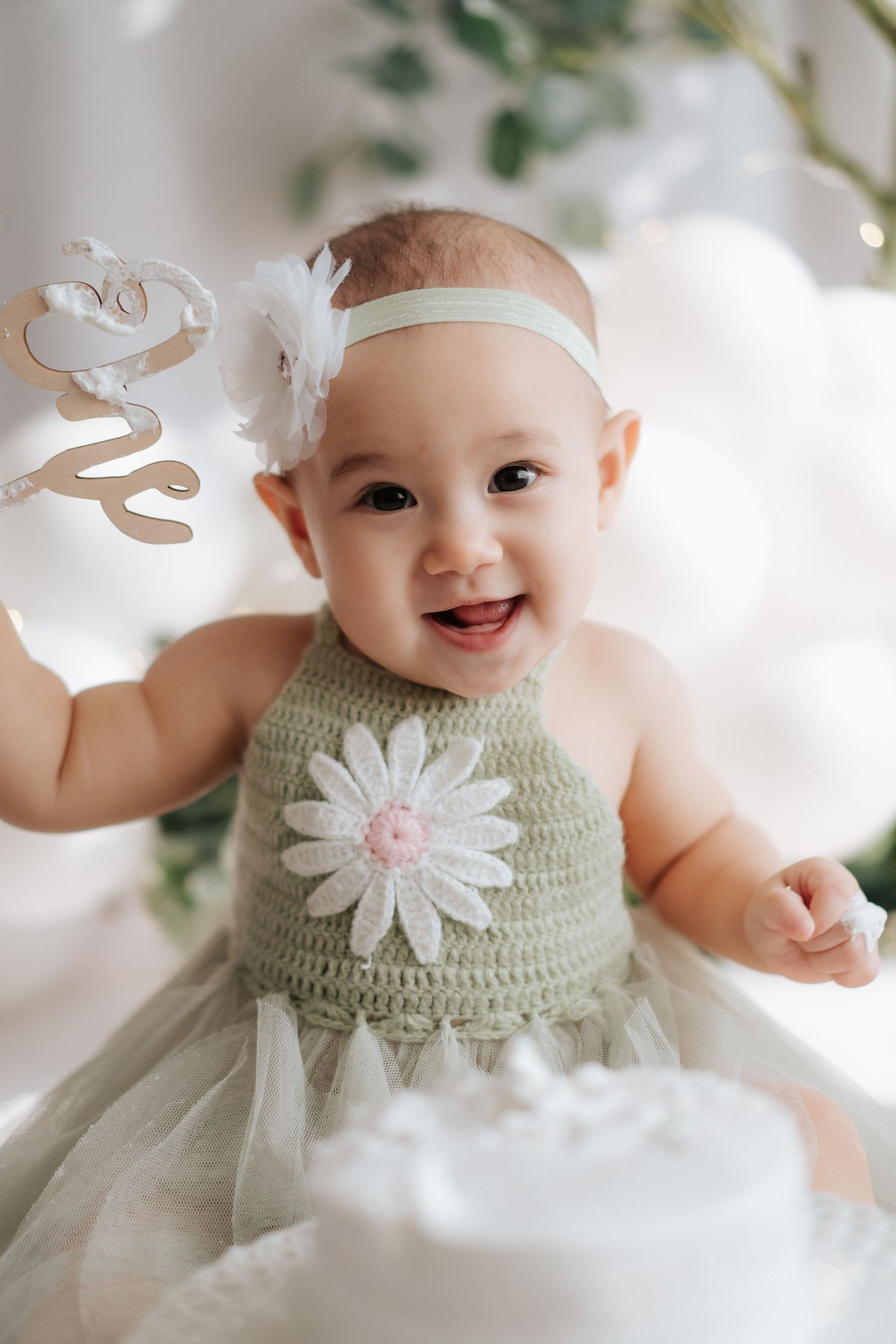 Baby in mint green dress with white daisy detail and tulle skirt smiles joyfully in bright setting.身穿薄荷綠連身裙,裙擺飾有白色雛菊圖案和薄紗裙擺的嬰兒,在明亮的背景下露出燦爛的笑