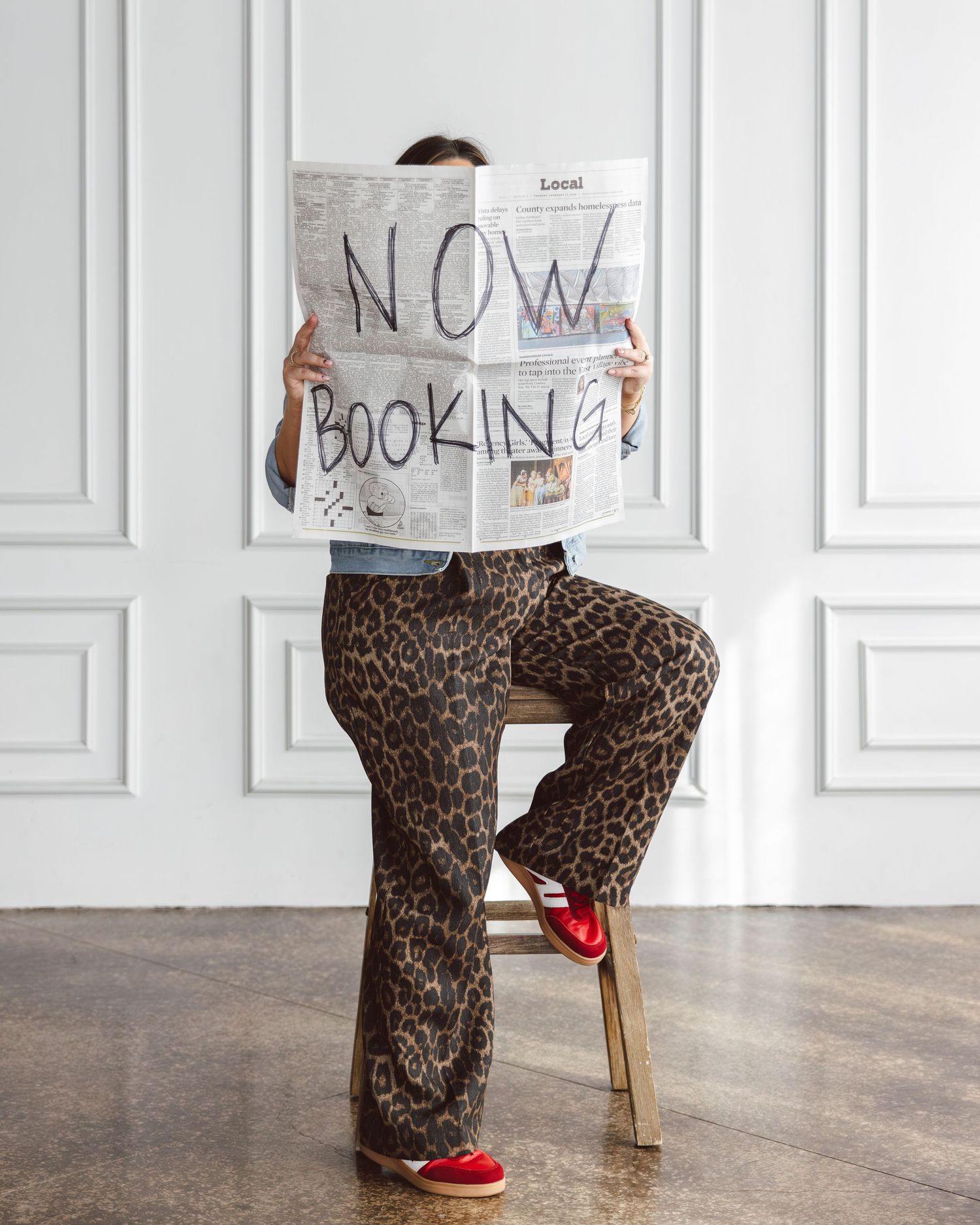 Person in leopard print pajamas and red slippers sits reading newspaper in white minimalist room.