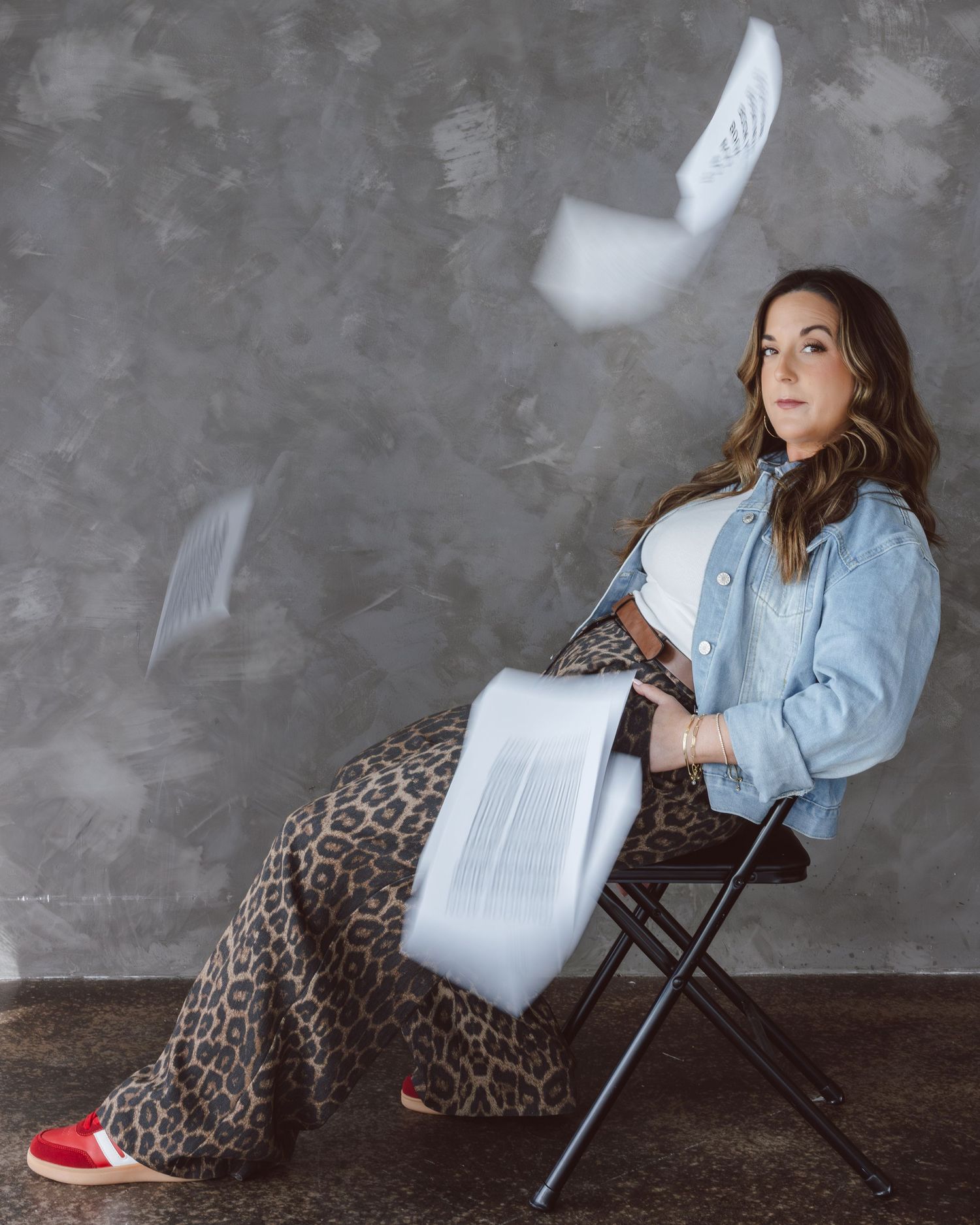A person in a denim jacket and leopard print skirt sits casually on a folding chair against a gray concrete wall.
