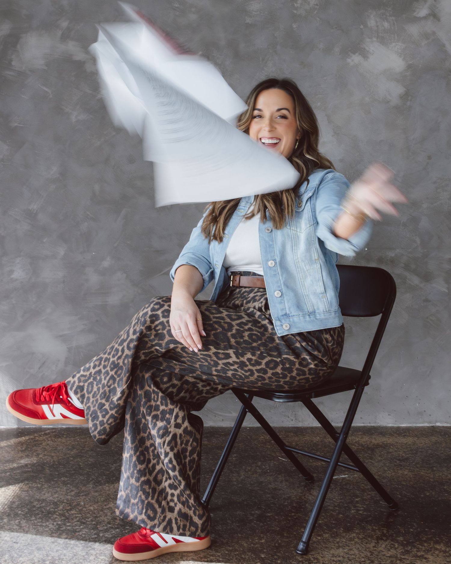 Woman in leopard print pants and denim jacket poses playfully on chair against gray wall.
