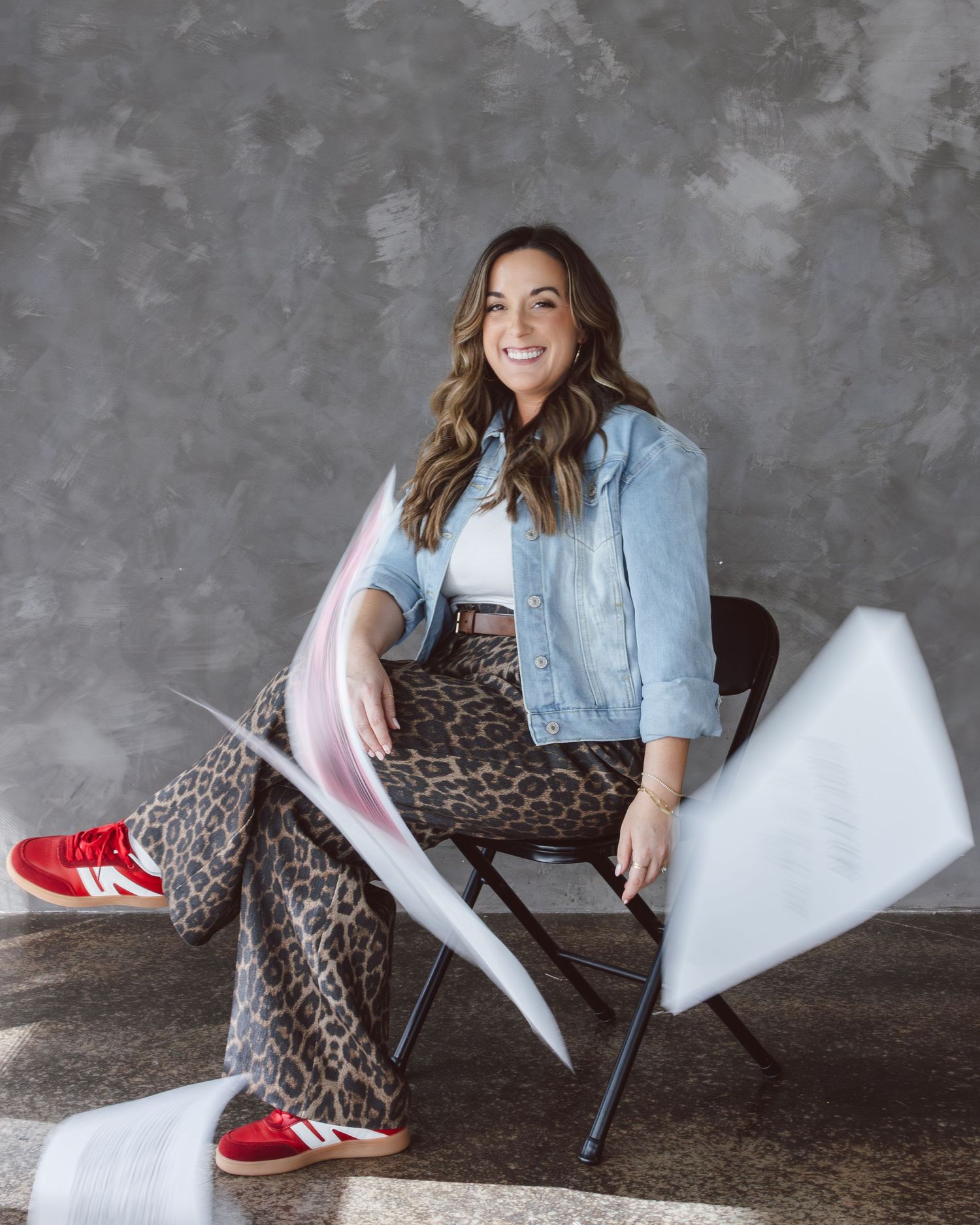Stylish casual outfit featuring leopard print pants, denim jacket, white top and red sneakers against textured grey wall.