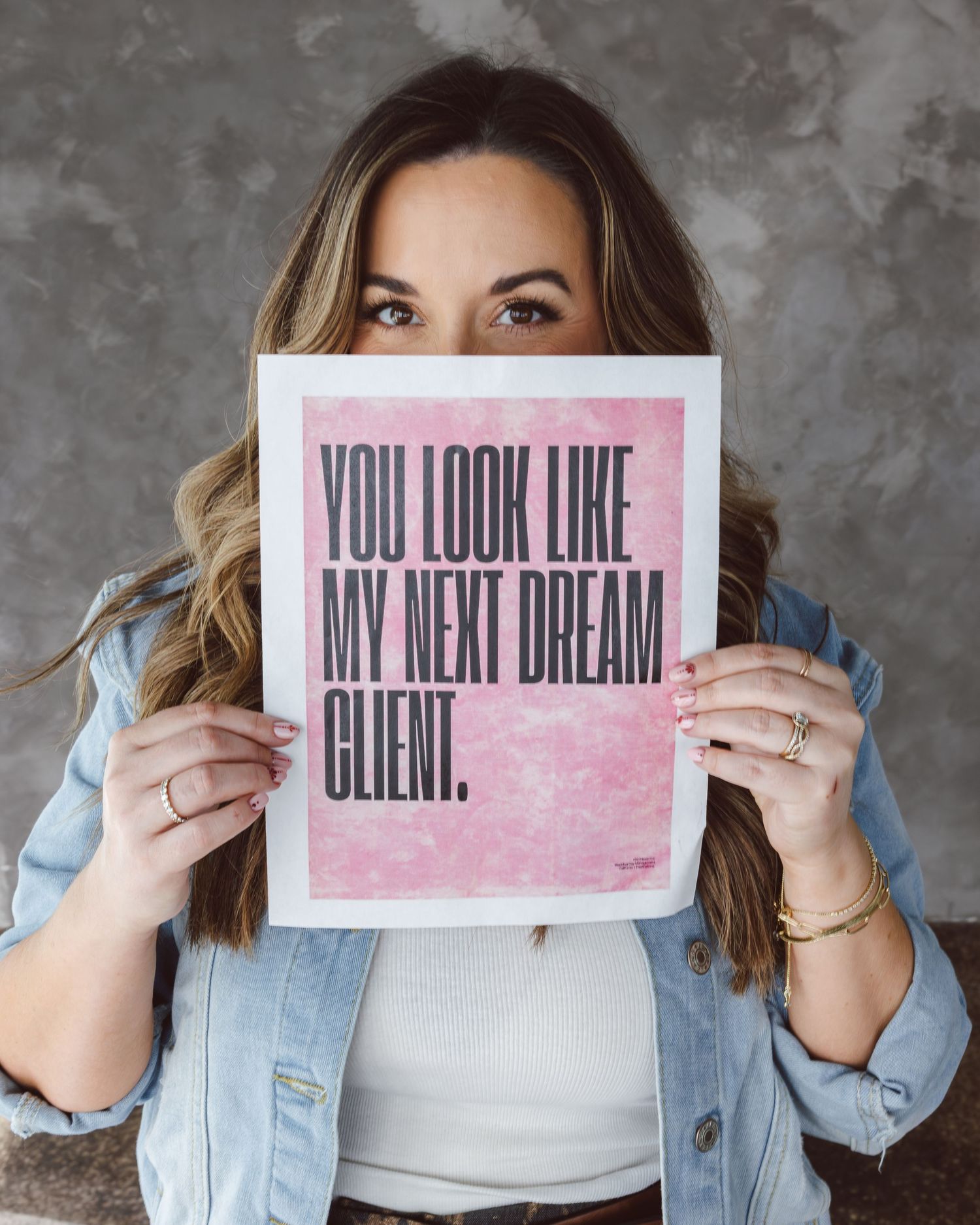 Person in denim jacket holds pink sign with text 'You look like my next dream client' against gray background.