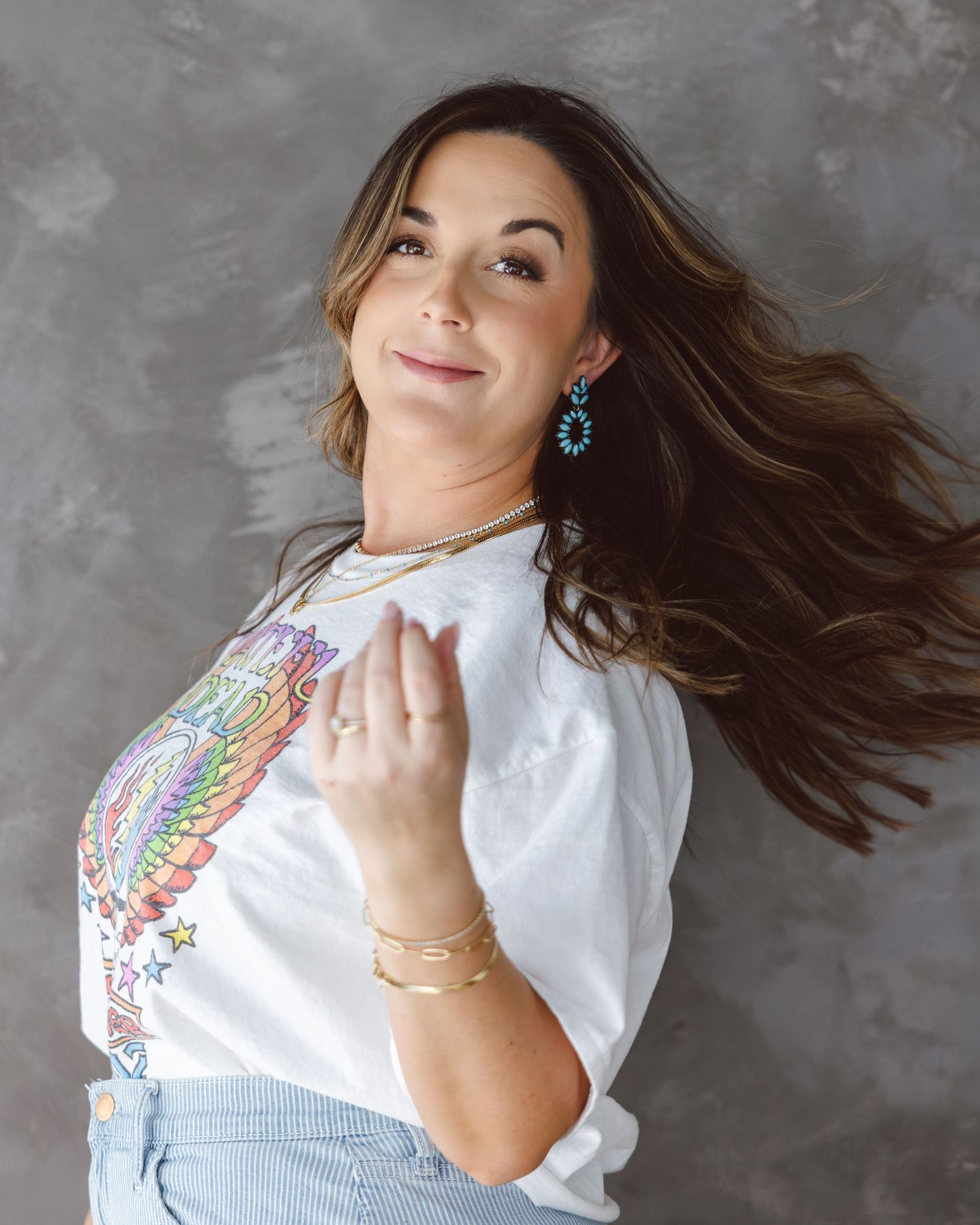 Woman in white t-shirt poses dynamically with flowing hair against textured gray backdrop.