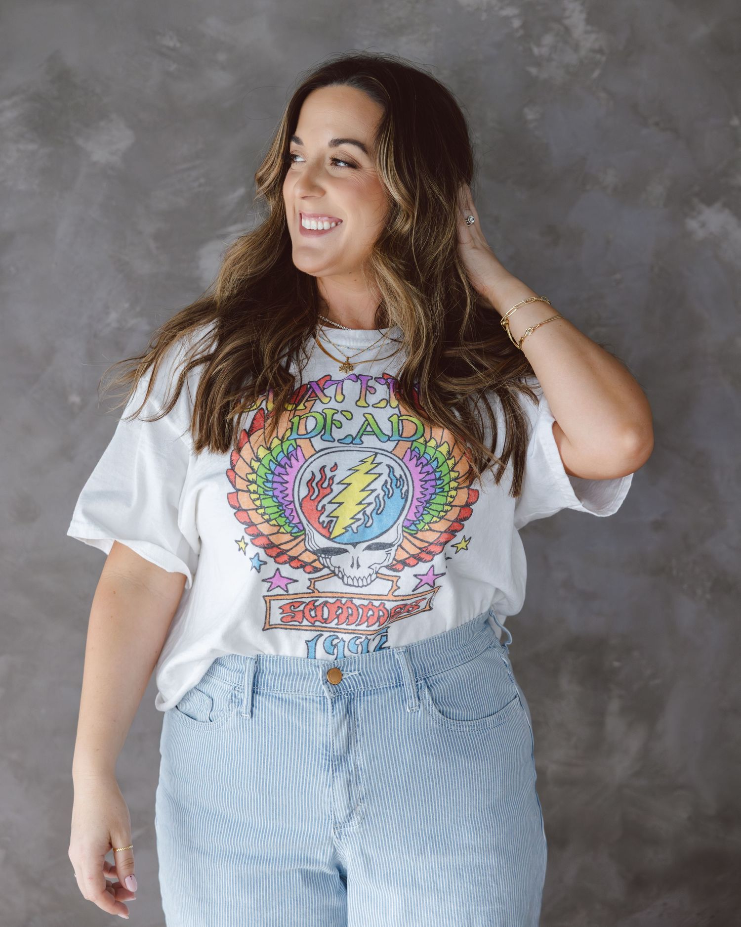 Smiling person wearing a white Grateful Dead graphic t-shirt and light blue jeans against a gray background.