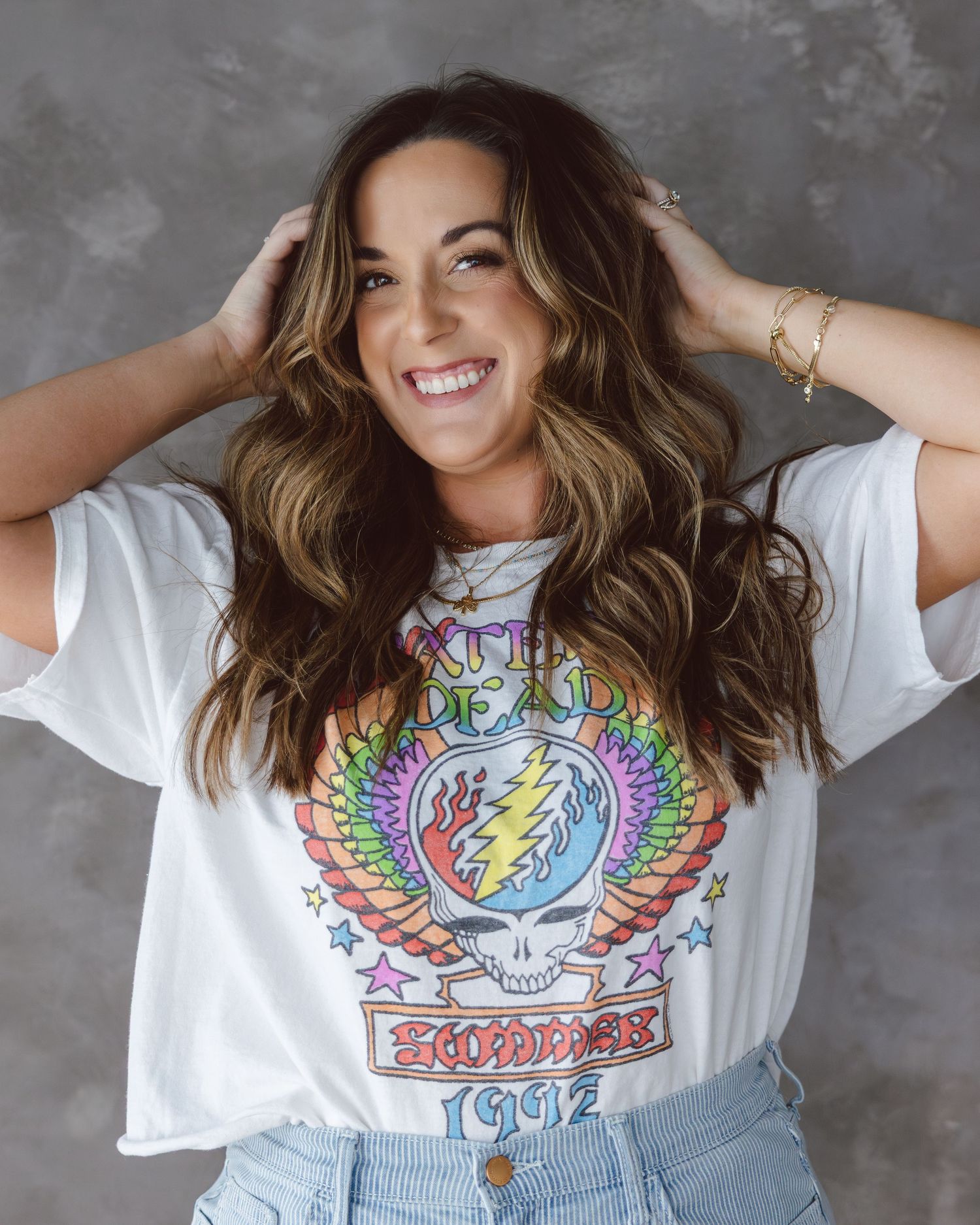 A person wearing a white t-shirt with a colorful winged skull graphic design smiles and poses against a gray background.