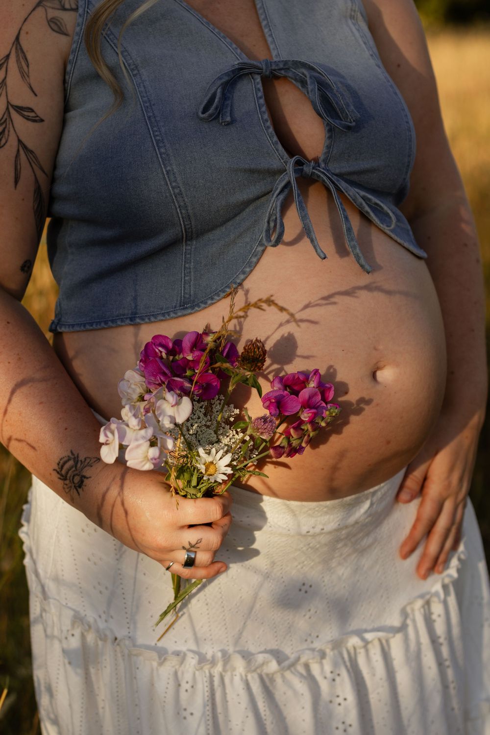 Pregnant woman in denim crop top and white skirt holding wildflowers bouquet outdoors in golden sunlight.
