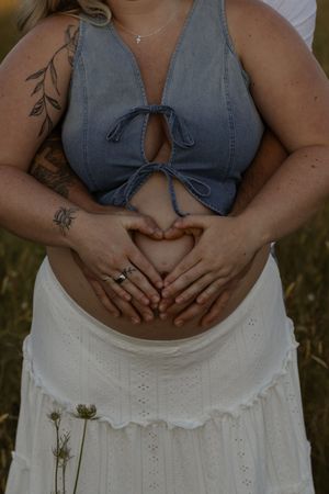 Pregnant couple making a heart shape with their hands on the baby bump, woman wearing a blue crop top and white skirt.
