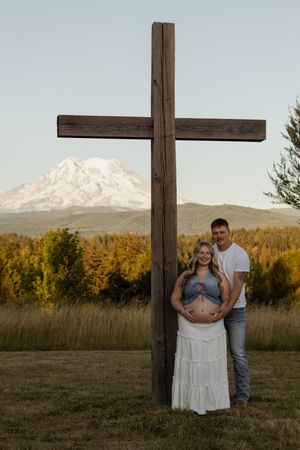 Pregnant couple poses by wooden cross with snow-capped Mount Rainier in background.