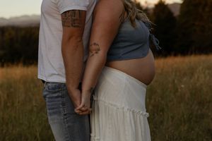 Pregnant couple holding hands in a golden field at sunset, showing baby bump in gray crop top and white skirt.