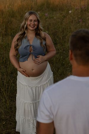 Smiling pregnant woman in denim crop top and white skirt holds baby bump in golden hour field.