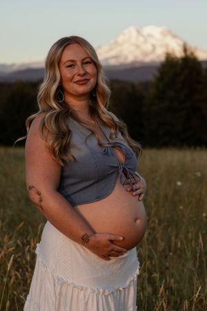 Pregnant woman in blue crop top and white skirt holding belly in field with snow-capped mount rainier backdrop.