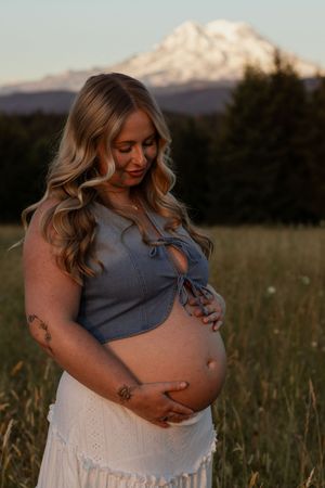 Pregnant woman in blue crop top and white skirt cradling baby bump in golden field with Mt. Rainier backdrop.