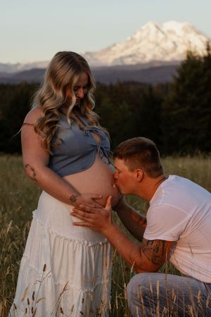A man kisses a pregnant woman's belly in a field with a snow-capped mount rainier backdrop at golden hour.