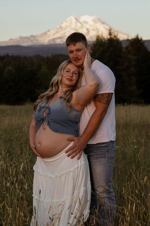 Pregnant couple embracing in a golden field with snow-capped Mount Rainier in the background during sunset.