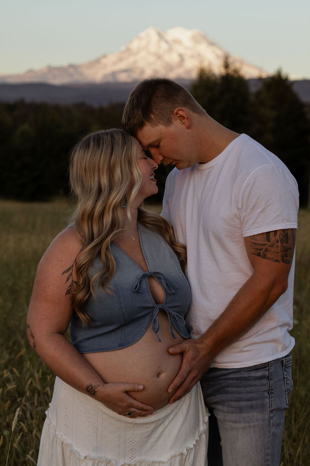 Expecting couple embracing in a field with Mount Rainier in the background at golden hour, woman in blue crop top.
