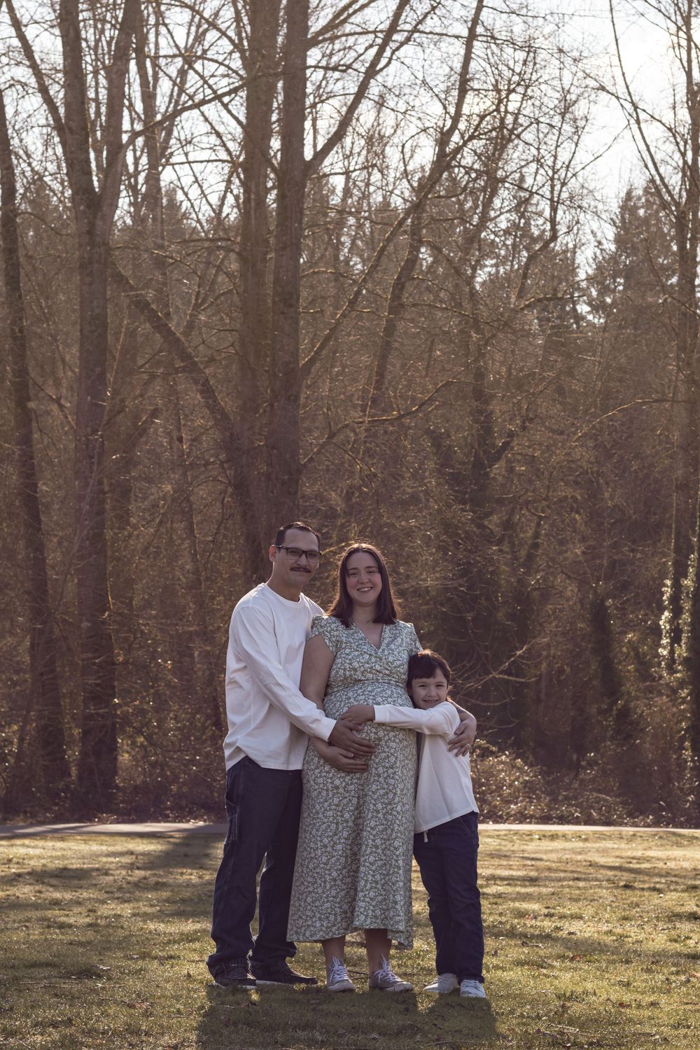 A family poses outdoors with a pregnant mother in a floral dress embraced by her partner and child.